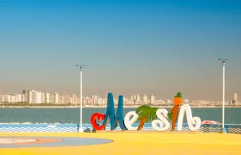 Mersin Merkez Gezilecek Yerler: 1 Günde 3 Etaplı Görülecek 15 Yer Mersin merkez sahil bandında yer alan Mersin şehir logosu, arkada masmavi Akdeniz ve şehrin modern silüeti.