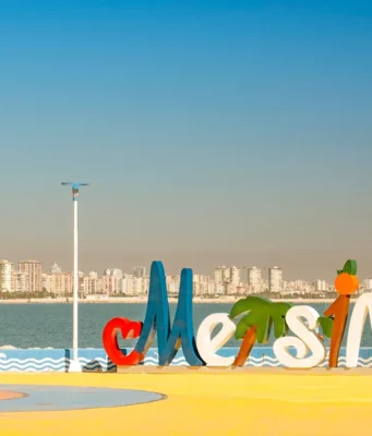 Mersin Merkez Gezilecek Yerler: 1 Günde 3 Etaplı Görülecek 15 Yer Mersin merkez sahil bandında yer alan Mersin şehir logosu, arkada masmavi Akdeniz ve şehrin modern silüeti.