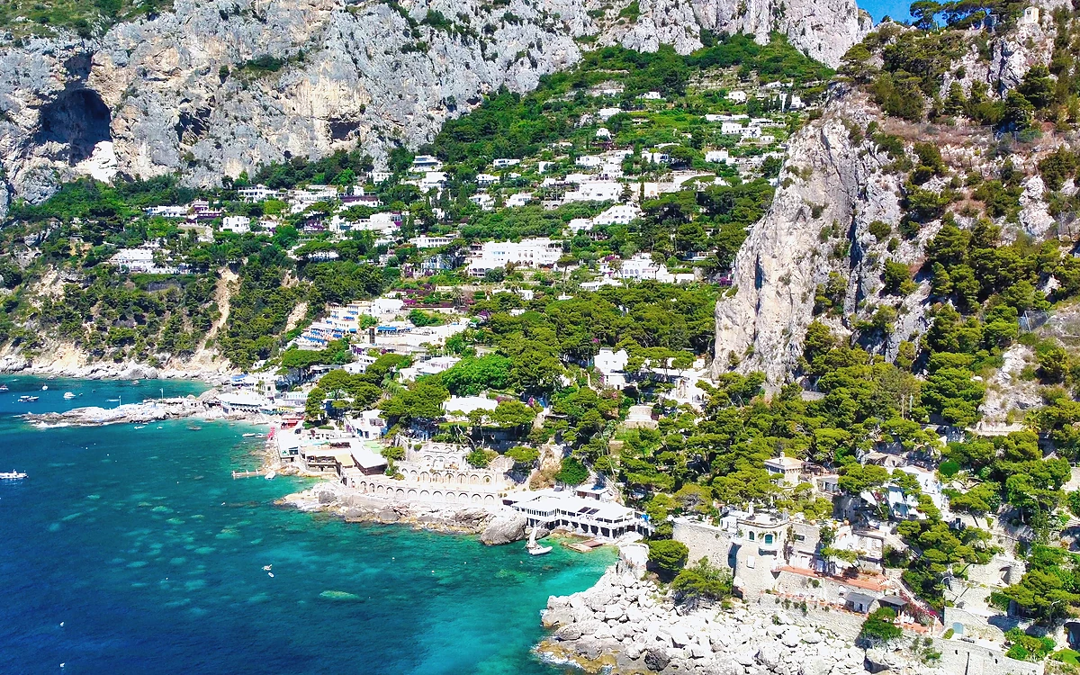 Capri Adası Marina Piccola koyu, kıyıdaki beyaz binalar ve sarp kayalık yamaçların panoramik görünümü.
