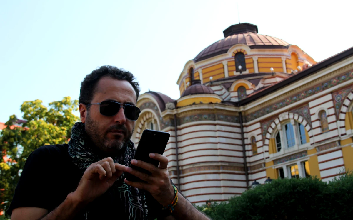 Sofya Merkez Kaplıcası binasının tarihi kubbeleri önünde telefonuna bakan güneş gözlüklü bir adam.