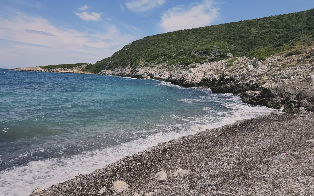 Güneşli bir günde Karaburun'da kayalık bir koyda, berrak turkuaz denizin ve yeşil tepelerin görüldüğü doğal bir manzara.