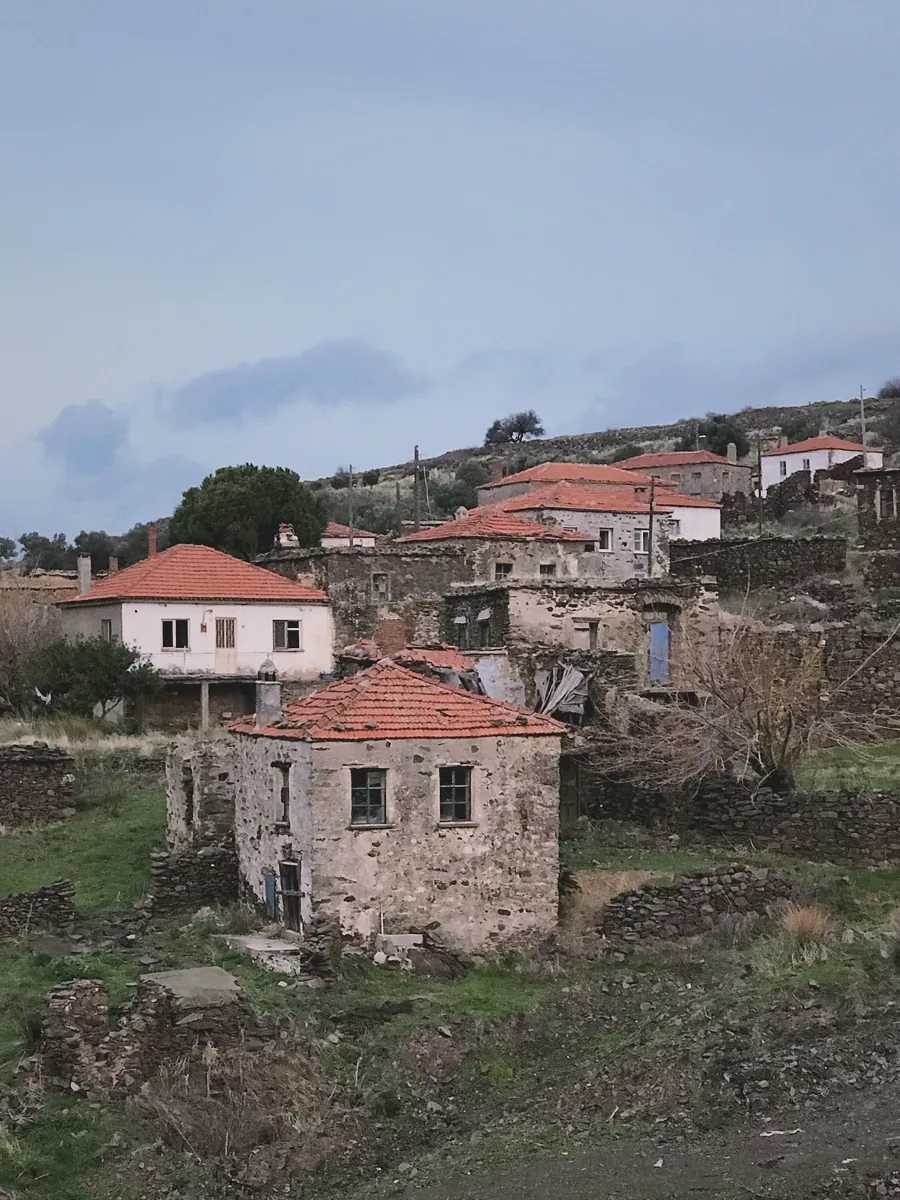 Bulutlu bir gökyüzünün altında, tepelik bir arazide yer alan, kırmızı kiremit çatılı eski taş evlerden oluşan terk edilmiş bir köy.